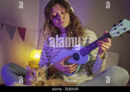 Junge Frau, die Ukulele übt, während sie Kopfhörer trägt, Musik in ihrem Schlafzimmer kreiert, zusammen mit einer einheimischen Ingwerkatze, die beobachtet, Freude findet und Stockfoto