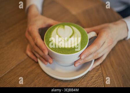 Frau hält eine weiße Tasse Matcha Latte mit wunderschöner Latte Art in Form eines Herzens, sitzt an einem Holztisch in einem Café und genießt einen Moment Stockfoto