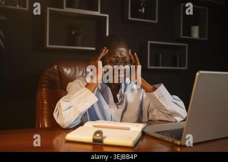 Junge afrikanische Geschäftsfrau, die mit schweren Kopfschmerzen zu kämpfen hat, während sie spät nachts im Büro arbeitet und ihre Schläfen massiert, um sie zu beruhigen Stockfoto