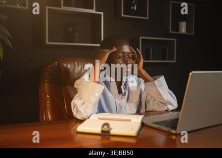 Professionelle Geschäftsfrau, die arbeitsbedingten Stress erlebt, Schläfen massiert, während sie bis spät an einem Laptop arbeitet, am Holzschreibtisch in schwachem l Stockfoto