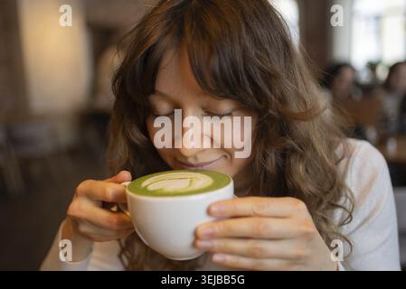Eine junge Frau mit lockigen Haaren genießt den Duft eines Matcha Latte in einem warmen und einladenden Café und genießt einen Moment der Ruhe inmitten des belebten Atmos Stockfoto