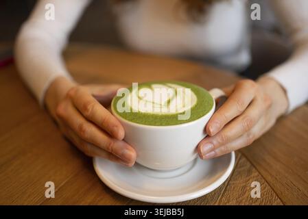 Frauenhände, die eine Tasse Matcha Latte mit herzförmigem Design in cremigem Schaum auf einem Holztisch liegen und ein warmes, gemütliches, gem Stockfoto