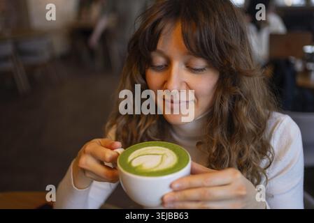 Junge Frau, die in einem gemütlichen Café einen warmen Matcha-Latte mit komplizierter Blattkunst genießt, genießt die friedliche Atmosphäre und die reichen, erdigen Aromen des Stockfoto