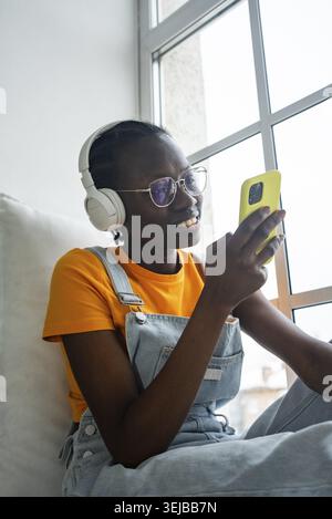 afroamerikanerin genießt Ausfallzeiten, trägt Kopfhörer und benutzt ein Smartphone. Am Fenster sitzt sie entspannt und zufrieden und nimmt einen an Stockfoto