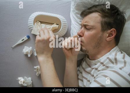 Kranker Mann, der im Bett liegt Husten und ein Gewebe aus einer Taschentüchtel nimmt, mit einem Thermometer und gebrauchten Geweben in der Nähe, leidet an Erkältung oder Grippe-Symptomen Stockfoto