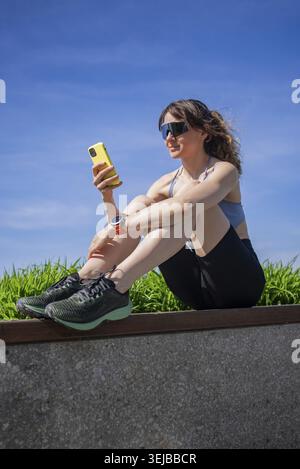 Die junge Sportlerin mit Sonnenbrille und Smartwatch nutzt nach dem Training im Freien eine Smartphone-App und sitzt auf einer niedrigen Wand mit grünem Gras unter bl Stockfoto