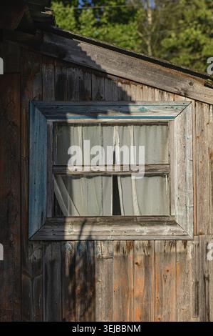 Nahaufnahme eines rustikalen Holzfensters mit weißen Vorhängen, eingerahmt von verwittertem Holz und beleuchtet von warmem Sonnenlicht. Das Bild erfasst ländliche CH Stockfoto