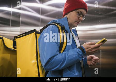 Der Liefermann mit rotem Wollhut und blauer Jacke überprüft sein Smartphone, während er einen gelb isolierten Thermorucksack für die Lieferung von Lebensmitteln trägt Stockfoto