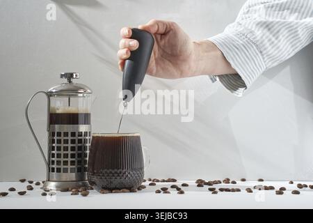 Verwenden Sie einen Handmilchaufschäumer, um frisch gebrühten schwarzen Kaffee in einer Tasse aus strukturiertem Glas neben einer französischen Metallpresse auf einer weißen Oberfläche zu mischen Stockfoto