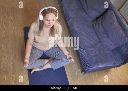 Reife weiße Frau übt zu Hause Meditation mit Kopfhörern, die auf dem Boden in der Nähe der Couch sitzen Stockfoto