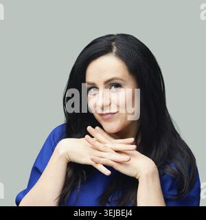 Lächelnde weiße Erwachsene mit langen schwarzen Haaren und blauem Hemd auf grauem Hintergrund Stockfoto