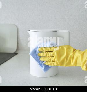 Person, die den weißen Wasserkocher mit gelben Handschuhen und blauem Tuch auf der Küchenzeile reinigt Stockfoto