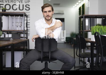 Junger Geschäftsmann mit ernsthaftem Ausdruck, der auf die Kamera blickt, während er hinten an vorne auf einem Stuhl im Büro sitzt Stockfoto