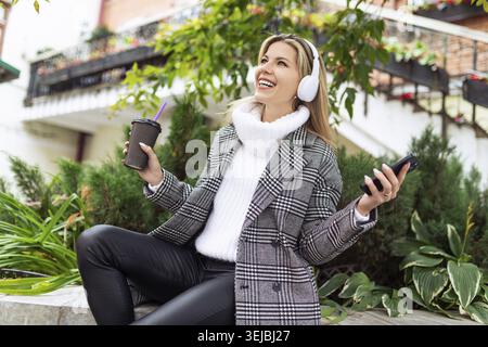 Eine erwachsene Frau genießt es, Musik in Kopfhörern im Hintergrund eines Stadtparks zu hören Stockfoto