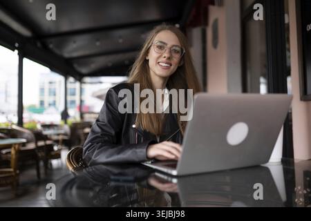 Die junge, stilvolle Frau sitzt auf der Treppe neben einem beliebten Café bei einer Tasse Kaffee und schaut sich ein Mobiltelefon an Stockfoto