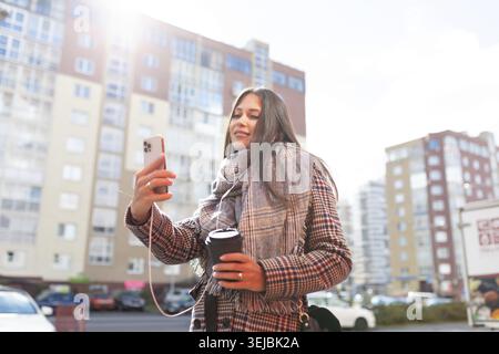 Stilvolle junge Frau, die mit einer Tasse Kaffee geht und ein Selfie in den Kopfhörern macht Stockfoto