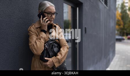 Eine ältere Frau, die am Eingang des Büros mit einem Mobiltelefon spricht Stockfoto