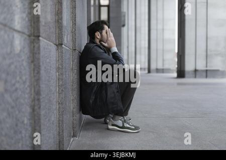 Ein Mann in einem deprimierten Zustand sitzt zurück an der Wand und bedeckt seine Hände mit seinen Handflächen Stockfoto