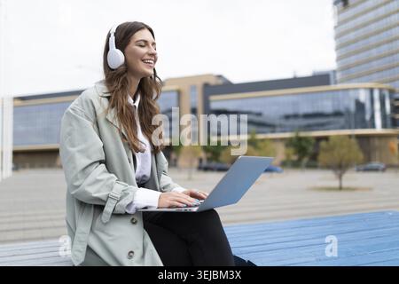 Freiberuflich tätige Frau mit Kopfhörern, die vor dem Hintergrund des Büros an einem Laptop arbeitet Stockfoto