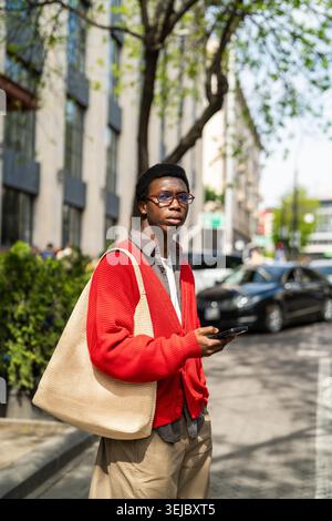 Fokussierter afroamerikanischer Student, der Smartphone in der Hand hält, auf der Straße steht und auf ein Taxi wartet. Stockfoto