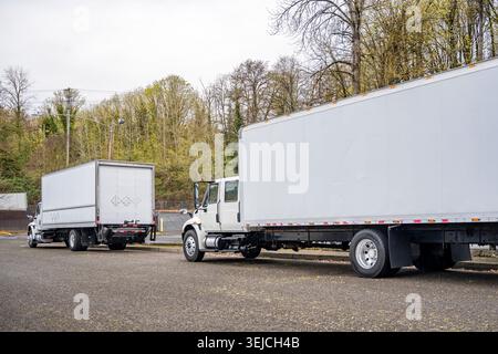 Kompakte, kompakte Sattelauflieger mit weißem Tagfahrerhaus in Industriequalität und langen Kastenanhängern auf der grünen Straße der Stadt und warten auf den nächsten Einheimischen Stockfoto