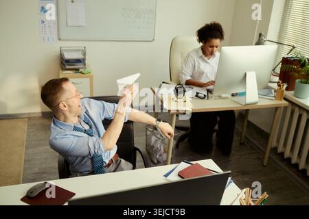 Hochwinkelansicht eines kaukasischen Geschäftsmannes, der Papierflugzeuge ins Büro wirft, während schwarze Geschäftsfrau am Desktop-Computer arbeitet und Ablenkung am Arbeitsplatz für HR-Inhalte zeigt Stockfoto