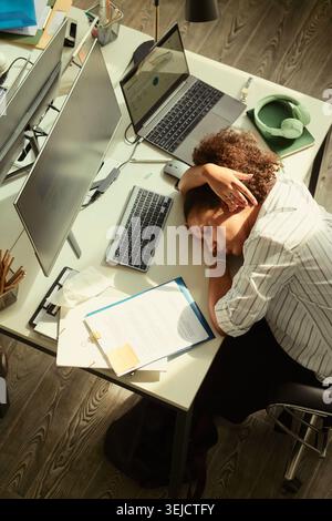 Hochwinkelansicht einer jungen, erwachsenen afroamerikanischen Geschäftsfrau, die auf dem Schreibtisch neben Laptop und Monitor steht und Arbeitserschöpfung und Burnout zeigt. Nützlich für Stressmanagement- oder Überarbeitskampagnen Stockfoto