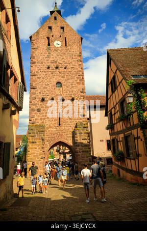 Riquewihr, Frankreich - 12.08.2023: Alter mittelalterlicher Glockenturm erhebt sich über einer engen Fußgängerzone mit Fachwerkhäusern und Wanderern Stockfoto