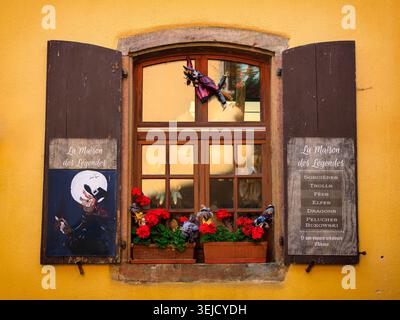 Riquewihr, Frankreich - 12.08.2023: Bezaubernde Schaufenster mit roten Geranien, dunklen Fensterläden und mythischen Texten auf einer gelben Fassade Stockfoto
