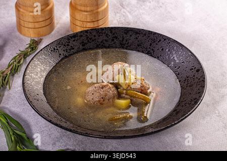 Herzhafte Fleischbällchensuppe mit Kartoffeln und grünen Bohnen in klarer Brühe. Serviert in einer strukturierten schwarzen Schüssel auf grauer Oberfläche mit frischen Kirschtomaten und Kräutern. Stockfoto