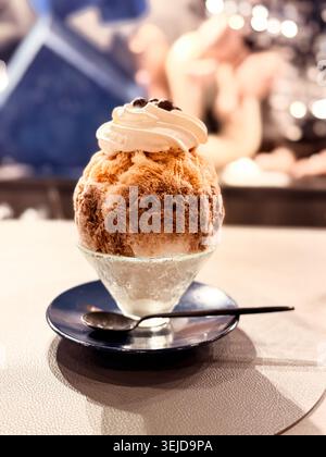 Ein wunderschön gefertigtes Kakigori-Eis mit Tiramisu-Geschmack in einer Glasschale auf einer dunklen Untertasse wird in einem Café in Akasaka, Tokio, mit einem traumhaften blauen Bokeh-Hintergrund fotografiert. Stockfoto