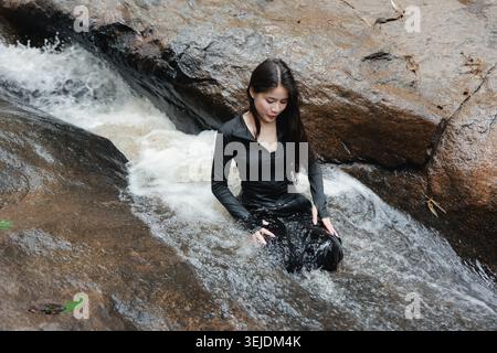 Junge asiatische Fotografin mit großer schwarzer Kamera, die im rauschenden Wasserfallbach sitzt und sich ruhig und konzentriert fühlt, umgeben von felsigem Naturland Stockfoto