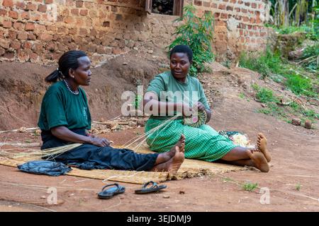 Gruppe afrikanischer Frauen, die auf dem Boden sitzen, lächeln und sich an traditionellem Handwerk des Webenens von Raffiakörben aus natürlichen Materialien beteiligen, umgeben von B Stockfoto