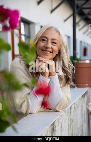Lächelnde, reife Frau, die sich auf den Balkon mit rosa Blumen lehnt Stockfoto