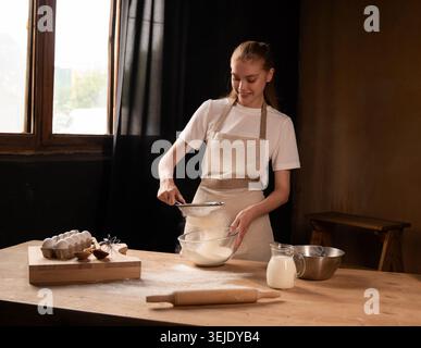 Frau siebt Mehl für hausgemachtes Backen, bereitet Teigzutaten in einer rustikalen Küche zu Stockfoto