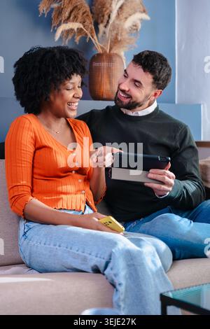Glückliches, multiethnisches Paar, das auf dem Sofa sitzt und auf dem Tablet-Bildschirm im Innenraum des Hauses schaut Stockfoto