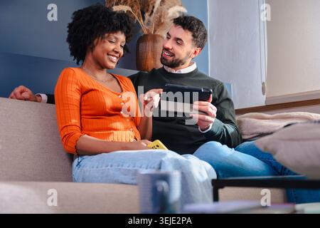 Glückliches, multiethnisches Paar, das auf dem Sofa sitzt und auf dem Tablet-Bildschirm im Innenraum des Hauses schaut Stockfoto