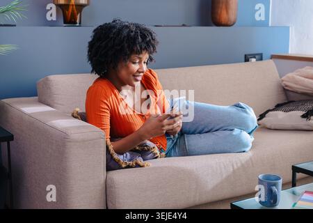 Glückliche junge Schwarze Frau, die auf der Couch mit Smartphone in einem hellen, gemütlichen Wohnzimmer liegt Stockfoto