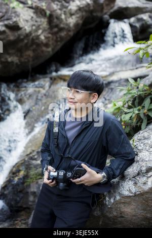 Junger asiatischer Fotograf mit friedlichem, fröhlichem Ausdruck, der die Kamera hält, die am natürlichen felsigen Wasserfall im grünen Wald steht und die Natur im Freien genießt Stockfoto
