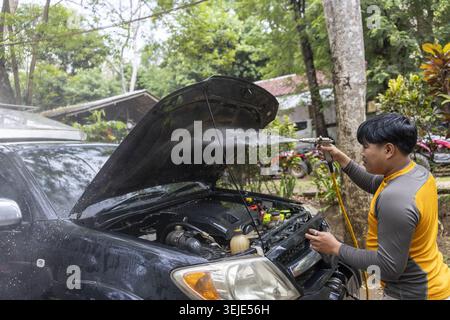 Fokussierter junger asiatischer Mann, der Autoreparatur und -Wartung wegen Pannenmotorproblemen durchführt. Dampf steigt aus dem Fahrzeug im Freien, während er an suv-Motor mit arbeitet Stockfoto