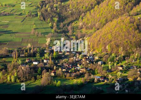 Ein Dorf in den Frühlingsbergen. Goldenes Licht taucht über sanfte grüne Hügel. Die Dächer des Dorfes schmiegen sich zwischen Feldern und Bäumen. Berge erheben sich dahinter, sanft im Schatten. Sonnenstrahlen erwärmen das Tal, sanft und hell Stockfoto