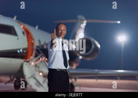Ein lächelnder Pilot in Uniform steht nachts auf dem Asphalt neben einem Privatjet und bietet einen Daumen nach oben. Professionell und selbstbewusst, vermittelt Luxusreisen, Business Aviation und einladenden Service. Stockfoto