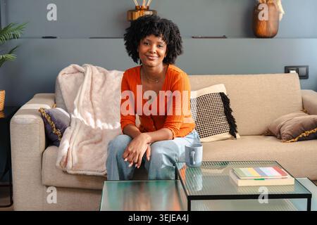 Lächelnde schwarze Frau in orangefarbenem Pullover, die auf einer Couch in einem gemütlichen Wohnraum sitzt Stockfoto