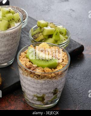 Zwei Gläser Chiasamenpudding, eines mit Kiwi-Würfel und das andere mit Müsli und Bananenchips, liegen auf einer dunklen Oberfläche Stockfoto