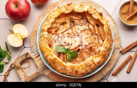 Hausgemachte Apfeltorte mit Mandelbelag auf einem Holzbrett, umgeben von frischen Äpfeln und Zimt Stockfoto
