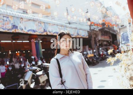 Nachdenkliche junge asiatische Frau, die auf der geschäftigen Stadtstraße in Asien steht. Lifestyle-Porträt einer schönen Reisenden, die einen urbanen Outdoor-Markt erforscht o Stockfoto