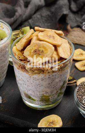 Zwei Gläser Chiasamenpudding, eines mit Kiwi-Würfel und das andere mit Müsli und Bananenchips, liegen auf einer dunklen Oberfläche Stockfoto