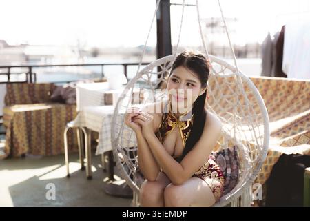 Nachdenkliche junge asiatische Frau entspannte sich und saß im Hängesessel draußen auf der sonnigen Terrasse. Zufriedene Person tagträumt mit nachdenklichem Blick während der friedlichen Zeit Stockfoto