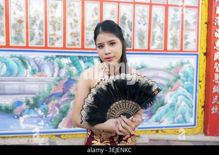 Schöne junge asiatische Frau in rotem traditionellem Kleid posiert anmutig mit schwarzem Handfächer und blickt in die Kamera mit selbstbewusstem Ausdruck in bunten Farben Stockfoto