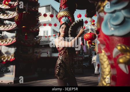 Elegante asiatische Frau im traditionellen Qipao-Kleid feiert freudig das chinesische Neujahr. Sie posiert mit Fächer draußen in festlicher roter Laterne und drago Stockfoto